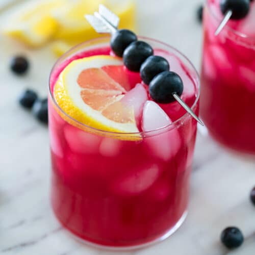 Overhead view of purple blueberry vodka lemonade with blueberry skewer and lemon wedge with second cocktail, blueberries and lemon wedges in background