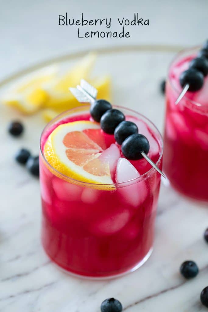 Glass of magenta colored blueberry vodka lemonade in a glass with eyes topped with a skewer of blueberries and a lemon wedge on a marble platter with blueberries and lemons in the background and "Blueberry Vodka Lemonade" in text at the top