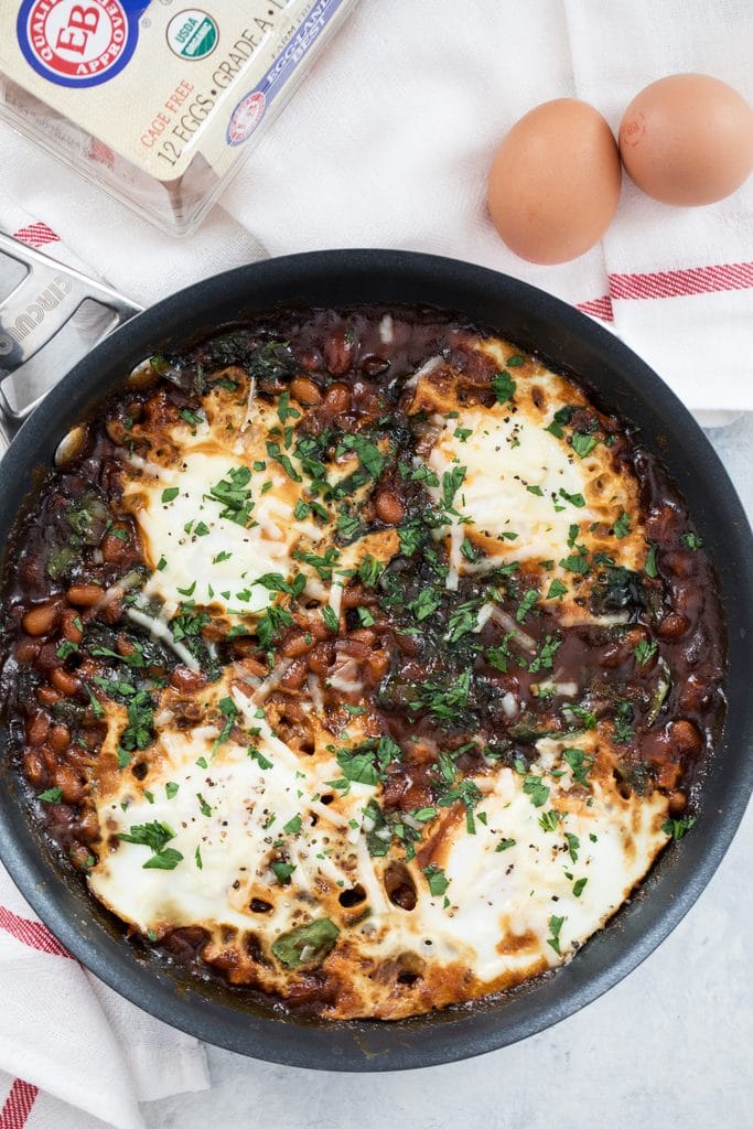 Overhead view of a skillet with Boston baked beans and eggs topped with parsley with whole eggs in the background