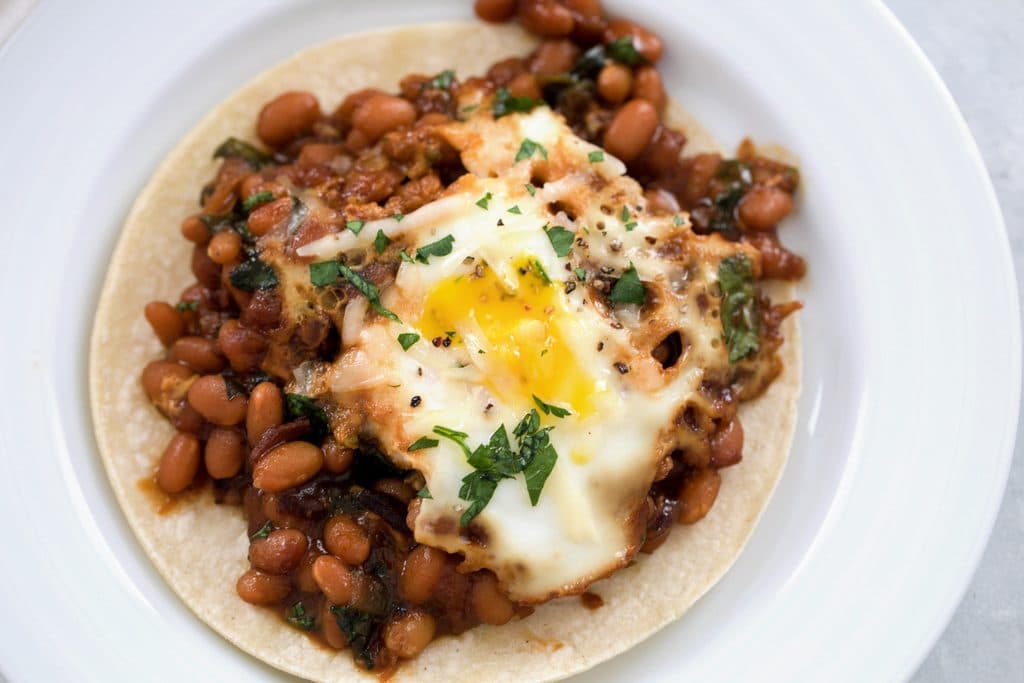 Overhead landscape view of a corn tortilla topped with Boston baked beans and eggs on a white plate