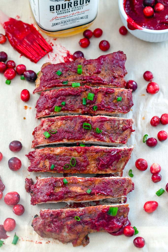 Overhead view of a rack of bourbon cranberry baby back ribs topped with green onions and surrounded by cranberries with a bowl of sauce, sauce brush, and bottle of bourbon in the background