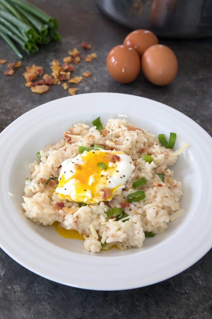 Breakfast risotto with bacon, poached egg and scallions with eggs, scallions, and crumbled bacon in background