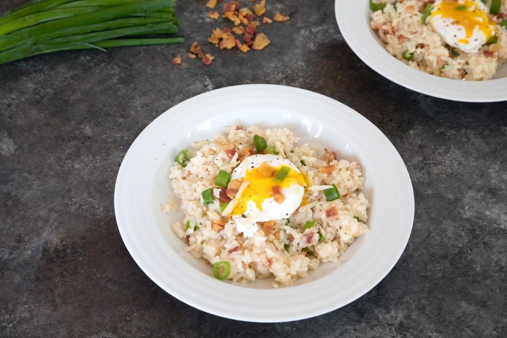 Landscape view of two bowls of breakfast risotto with runny poached eggs with scallions and crumbled bacon in background