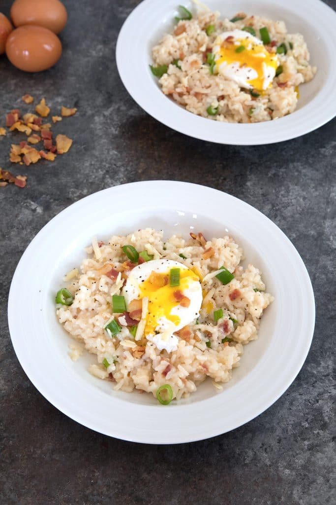 Overhead view of two bowls of breakfast risotto with poached eggs with crumbled bacon and eggs in background