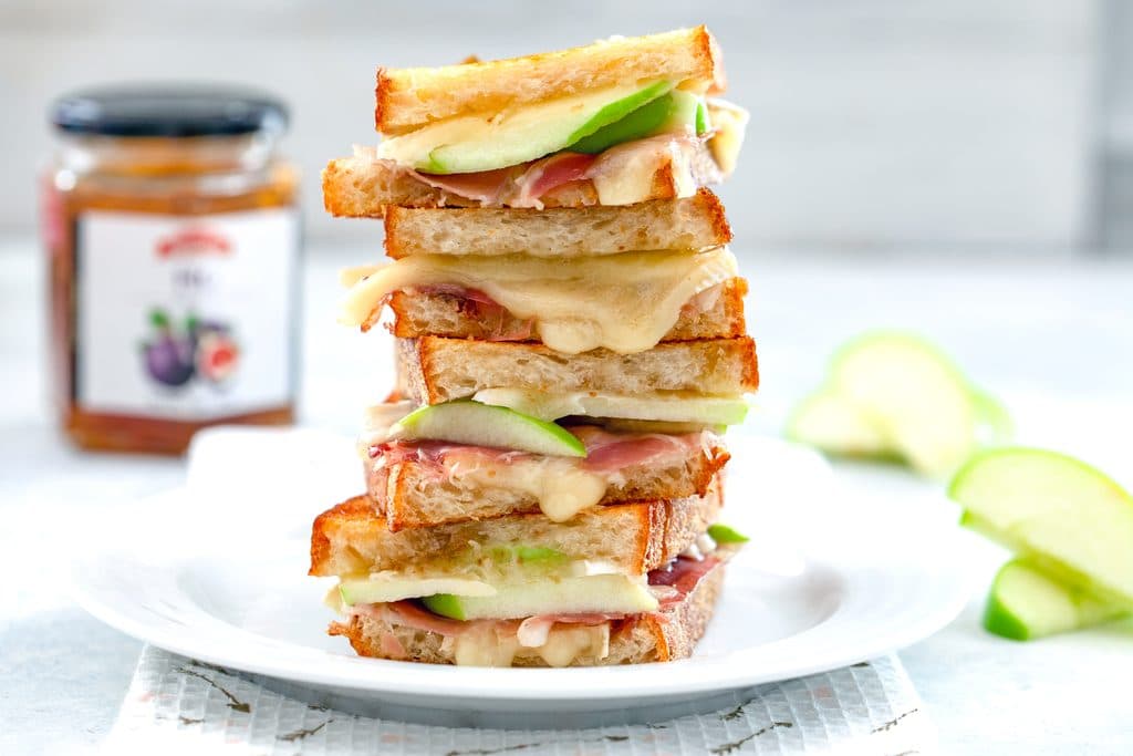 Landscape photo of four halves of a brie grilled cheese sandwich with green apple and prosciutto stacked on each other on a white plate with more apple and jar of fig spread in the background