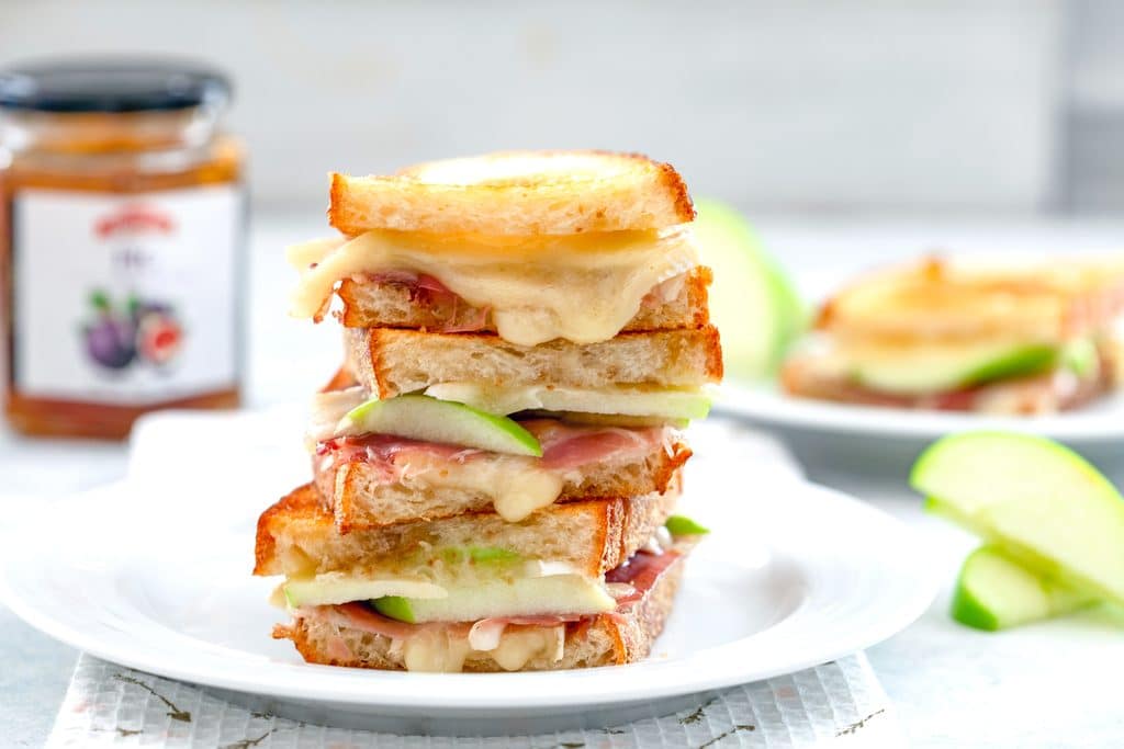 Landscape photo of three halves of a bri grilled cheese sandwich with green apples and prosciutto stacked on top of each other on a white plate with apples and jar of fig spread in the background