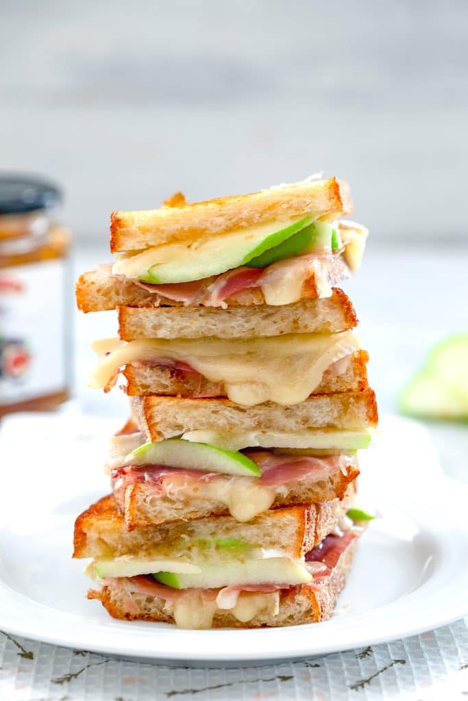 Four halves of a brie grilled cheese sandwich with fig spread, green apple, and prosciutto all stacked on each other on a white plate 