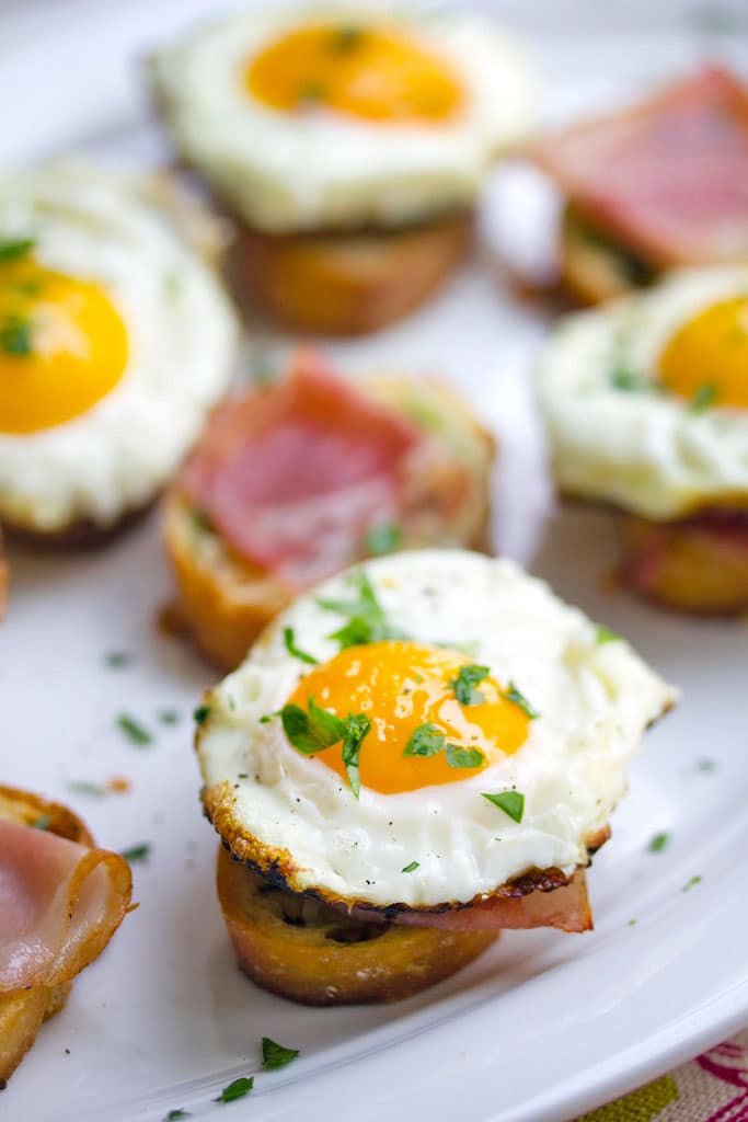 Overhead view of multiple brunch bruschetta with prosciutto and fried eggs with chopped parsley on a white platter