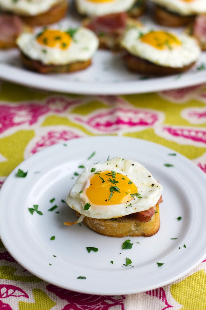 Head-on view of a brunch bruschetta with prosciutto, fried eggs, and parsley on a white plate with a platter filled with brunch bruschetta behind it