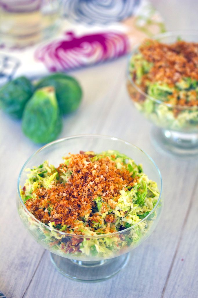 Head-on closeup view of bowl of brussels sprouts bacon caesar salad topped with panko with second bowl of salad, brussels sprouts