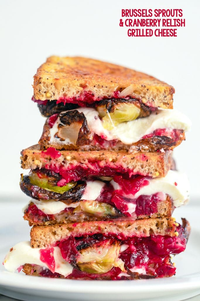 Head-on close-up view of three brussels sprouts and cranberry relish grilled cheese sandwich halves stacked on top of each other on a white plate with recipe title at top of image