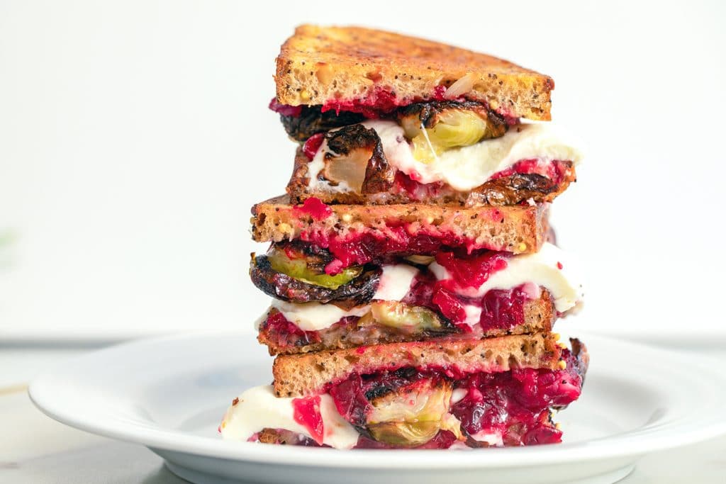 Landscape view of three halves of a brussels sprouts and cranberry relish grilled cheese sandwich stacked on top of each other on a white plate