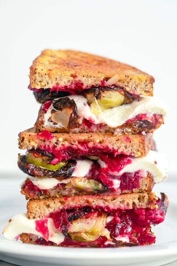 Head-on close-up view of three brussels sprouts and cranberry relish grilled cheese sandwich halves stacked on top of each other on a white plate