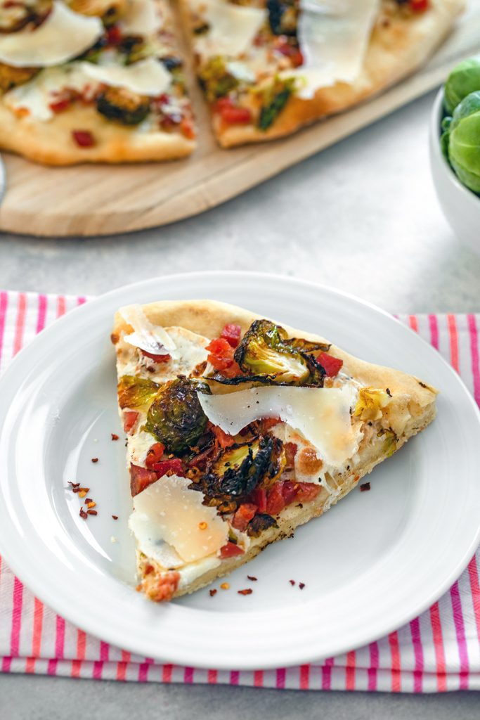 Overhead view of a slice of brussels sprouts flatbread with pancetta and shaved parmesan on a white plate with rest of pizza in background