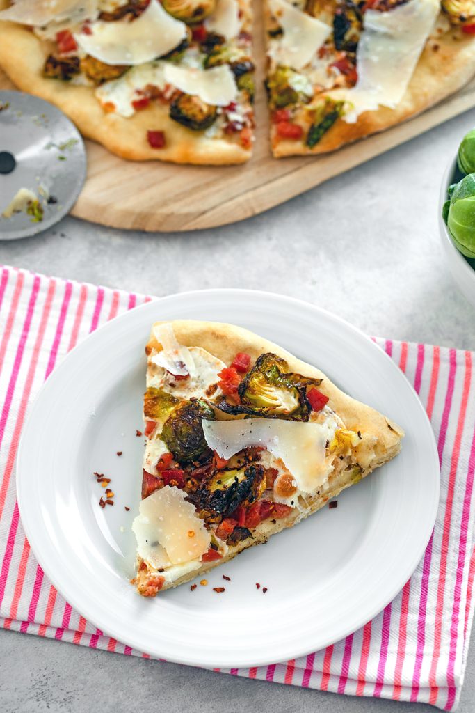 Overhead view of a slice of brussels sprouts flatbread with pancetta and parmesan on a white plate with rest of pizza and pizza cutter in background