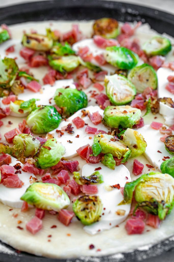 Overhead view of flatbread dough rolled out on pan with sliced brussels sprouts, pancetta, mozzarella, and red pepper flakes ready for the oven