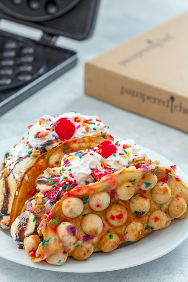 Head-on view of two bubble waffle banana split tacos with lots of toppings with Bubble Waffle Pan and Pampered Chef box in background