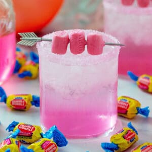 Head-on view of a pink bubblegum cocktail with gum garnish and Double Bubble gum all around with pink balloon and simple syrup in background