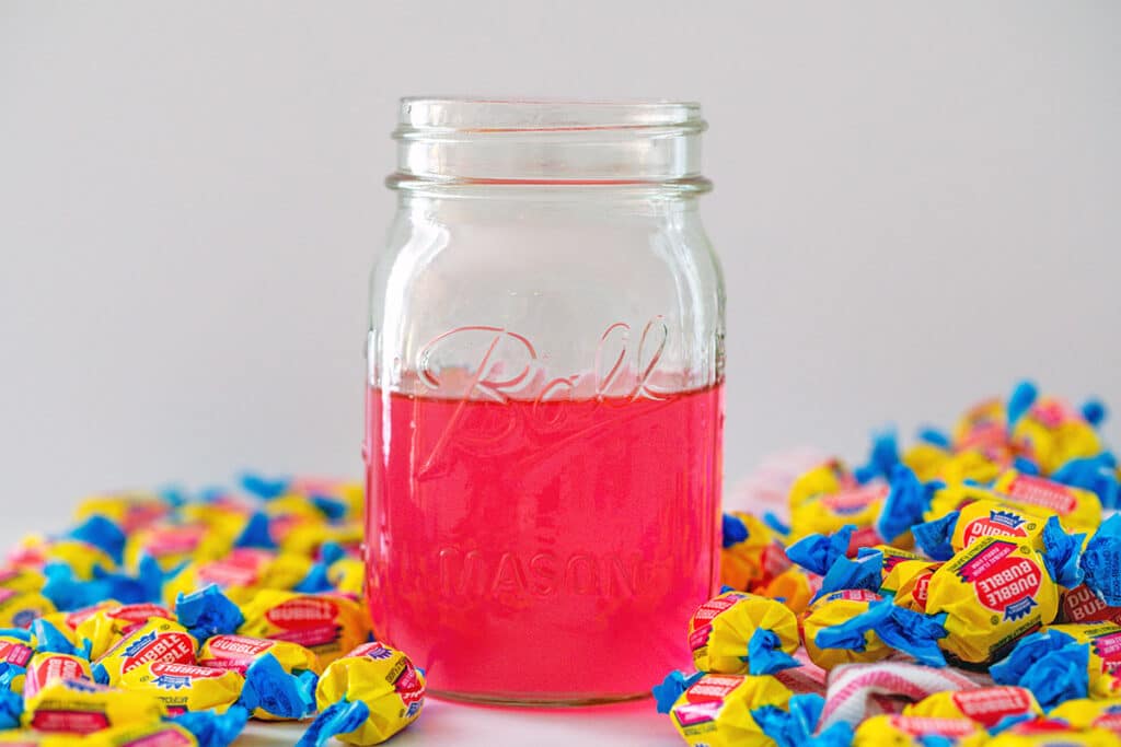 Landscape head-on view of a mason jar with pink bubblegum simple syrup with pieces of Double Bubble all around.