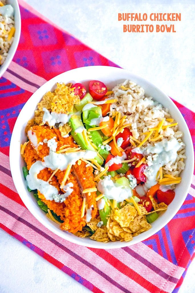 Overhead view of buffalo chicken burrito bowls with chicken avocado, tortilla chips, tomatoes, cheese, and rice, drizzled with blue cheese dressing and recipe title at top