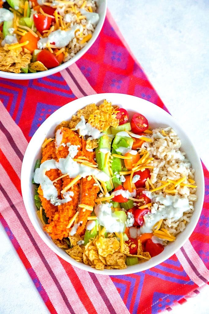 Overhead view of buffalo chicken burrito bowls with chicken avocado, tortilla chips, tomatoes, cheese, and rice, drizzled with blue cheese dressing with second salad bowl in background