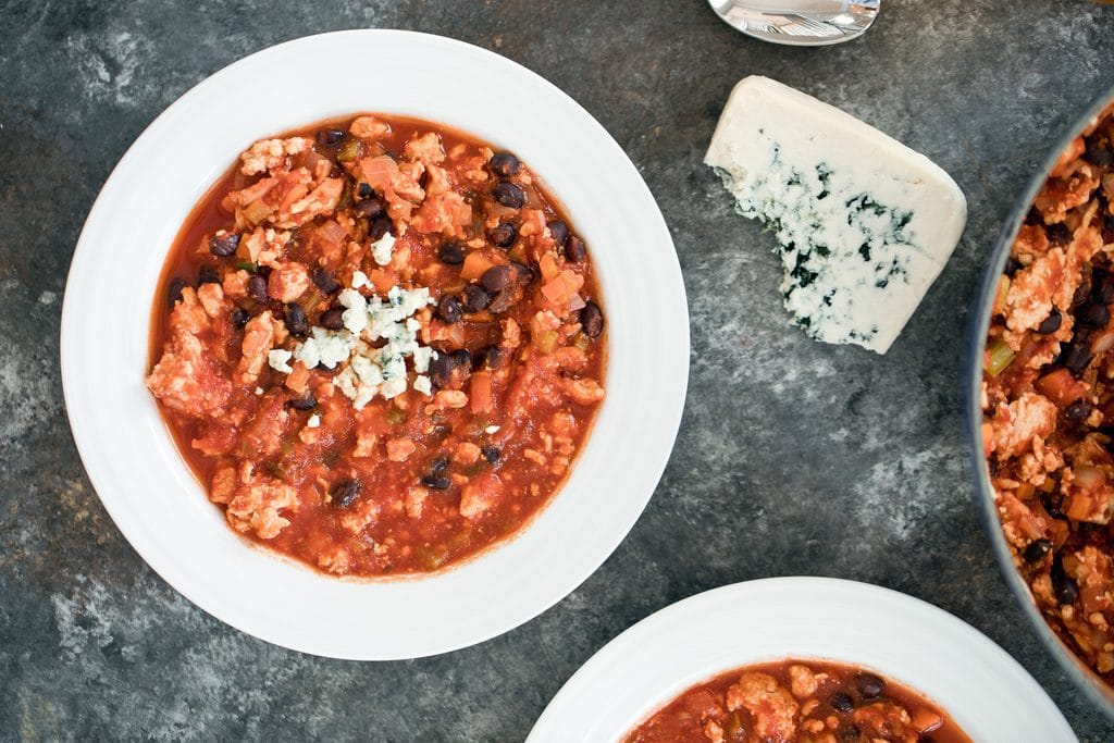 Landscape overhead view of bowl of buffalo chicken chili with blue cheese crumbled on top with second bowl of chili, pot of chili, and wedge of blue cheese in background