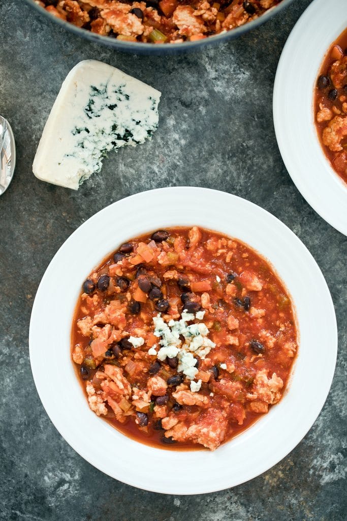 Overhead view of a bowl of buffalo chicken chili with blue cheese crumbled on top with wedge of blue cheese and second bowl of chili in the background