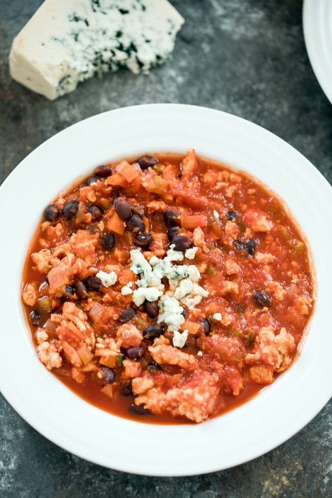 Closeup overhead view of bowl of buffalo chicken chili with blue cheese crumbled on top with wedge of blue cheese in the background