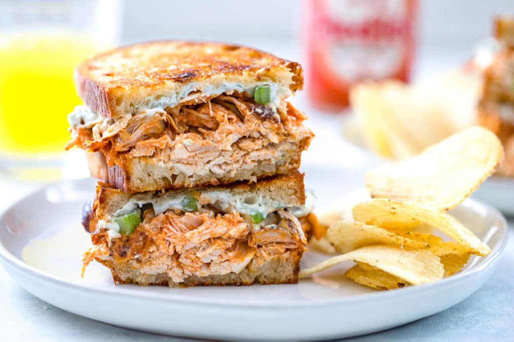 Landscape photo of two halves of buffalo chicken grilled cheese stacked on each other on gray plate with potato chips on plate and juice and Frank's Red Hot bottle in background.