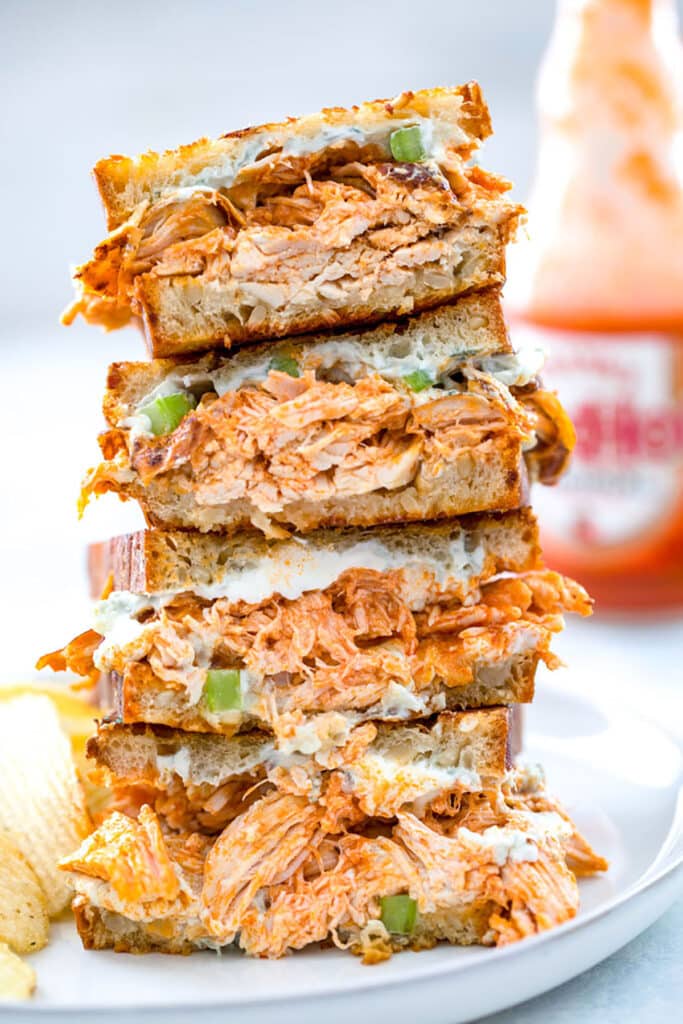 Four halves of buffalo chicken grilled cheese sandwich all stacked on each other on a gray plate with bottle of Frank's Red Hot in the background.