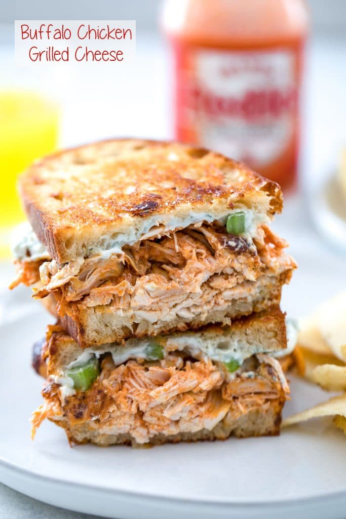 Two halves of a buffalo chicken grilled cheese sandwich stacked on each other on a gray plate with bottle of Frank's Red Hot in the background and "Buffalo Chicken Grilled Cheese" text at top