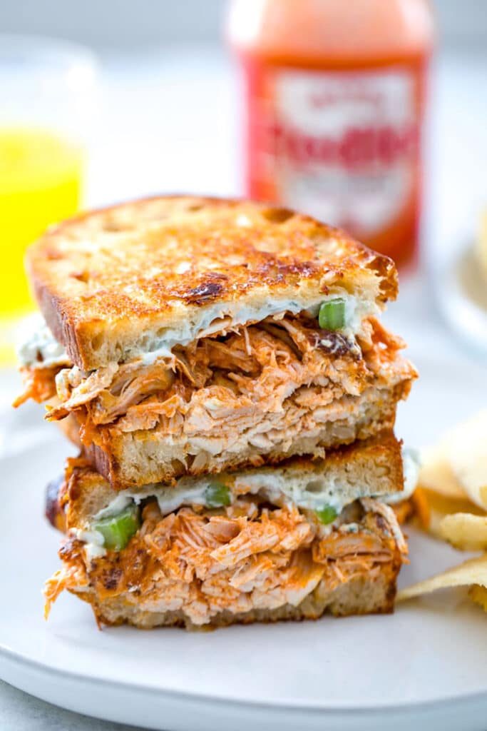 Two halves of a buffalo chicken grilled cheese sandwich stacked on each other on a gray plate with bottle of Frank's Red Hot in the background.