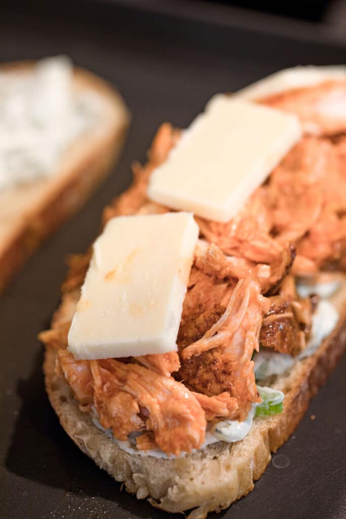 Bread with blue cheese spread, buffalo chicken, and cheese slices on griddle.