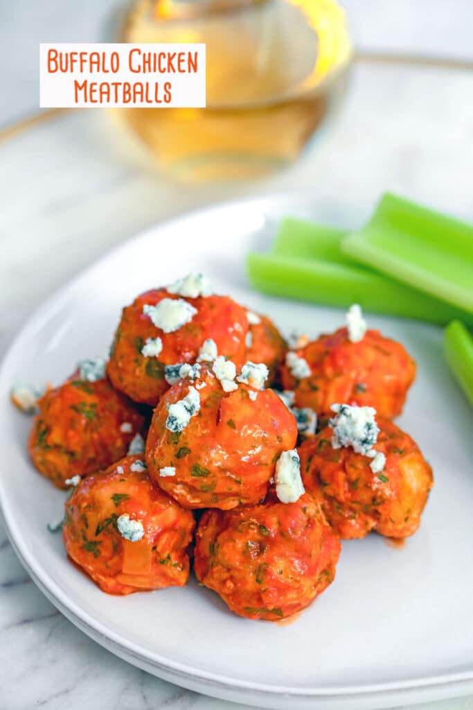 Overhead view of buffalo chicken meatballs topped with blue cheese and served with celery sticks with glass of white wine in the background and recipe title at top.