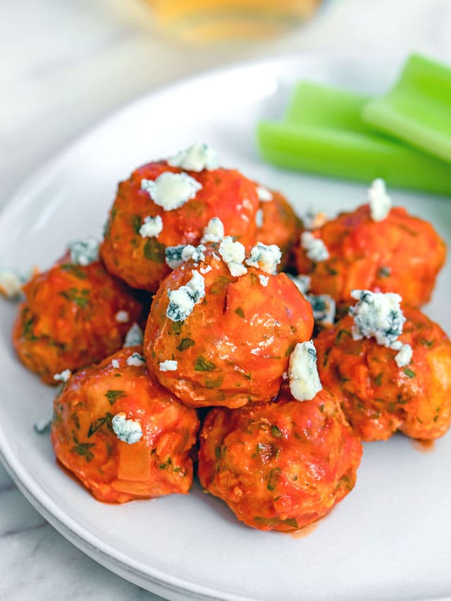 Overhead view of buffalo chicken meatballs topped with blue cheese and served with celery sticks with glass of white wine in the background.