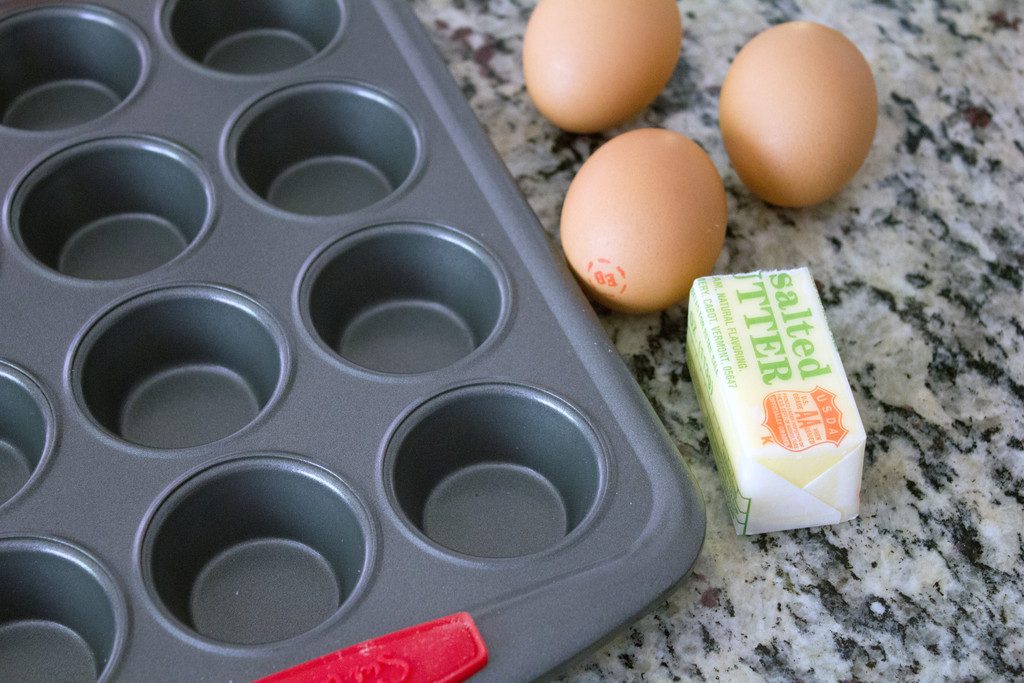 Overhead view of a mini cupcake pan with 3 eggs and a half stick of butter on a granite counter