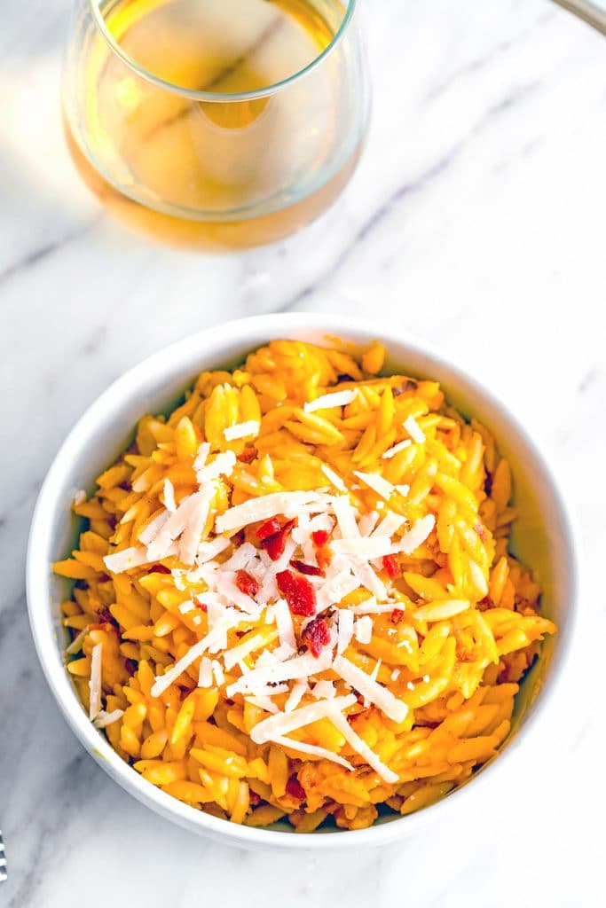 Overhead view of bowl of butternut squash orzo with grated parmesan and crumbled bacon on marble surface with glass of wine in the background