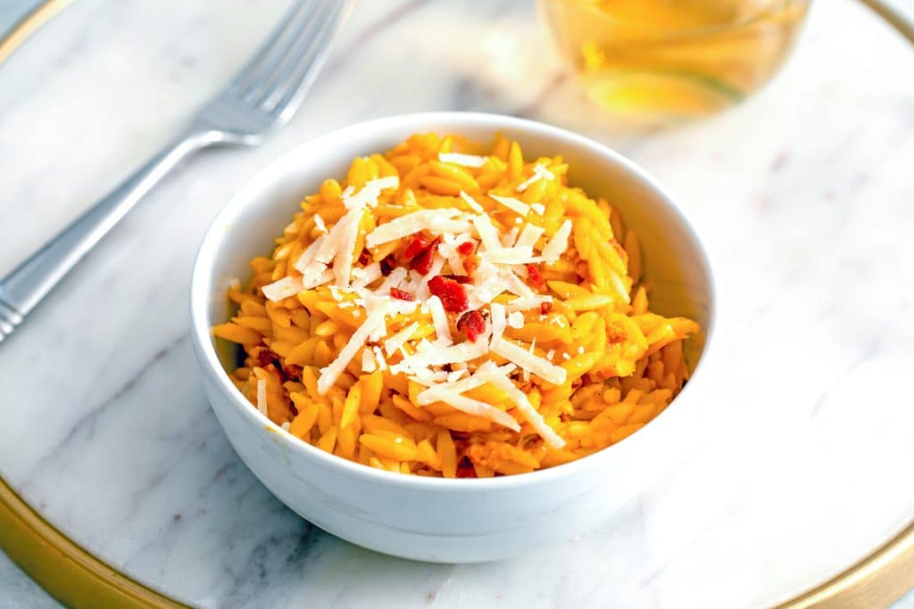 Landscape head-on view of butternut squash orzo in a white bowl topped with grated parmesan and crumbled bacon on marble surface with fork and glass of wine in background