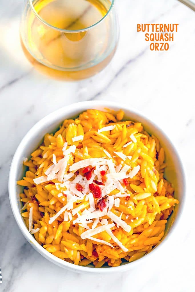 Overhead view of bowl of butternut squash orzo with grated parmesan and crumbled bacon on marble surface with glass of wine in the background and recipe title at top