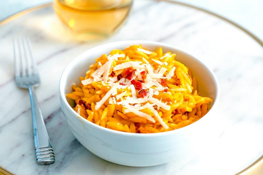 Head-on close up view of butternut squash orzo topped with parmesan cheese and crumbled bacon in a white bowl with fork and glass of white wine in background