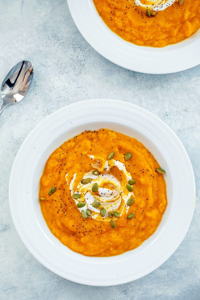 Overhead view from a distance of white bowl of butternut squash soup topped with sour cream and pumpkin seeds with spoon and second bowl of soup in background