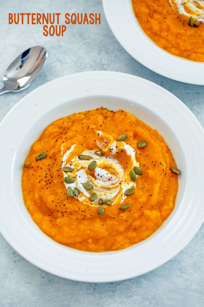 Overhead angled view of a white bowl of butternut squash soup topped with sour cream and pumpkin seeds with a spoon and second bowl of soup in the background and recipe title at top