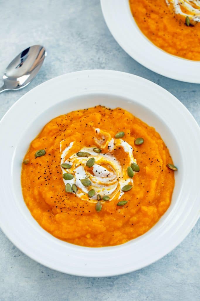 Overhead angled view of a white bowl of butternut squash soup topped with sour cream and pumpkin seeds with a spoon and second bowl of soup in the background