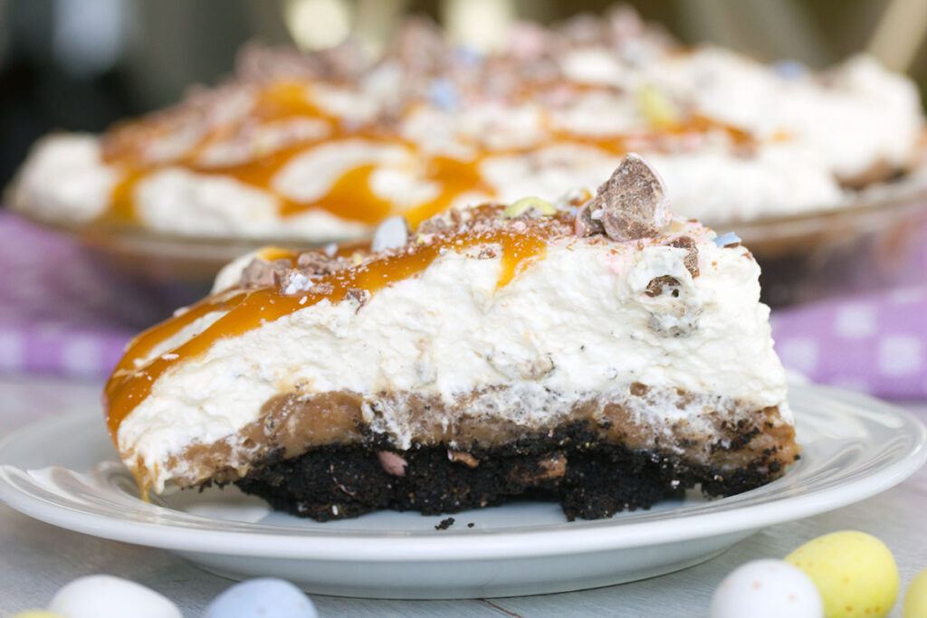 Landscape closeup view of a slice of Cadbury Egg pie showing layers of chocolate cookie crust, chocolate filling, and whipped cream topping topped with caramel and Cadbury Mini Eggs.