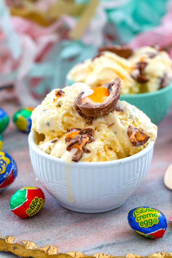 Head-on view of a white bowl of Cadbury Creme Egg ice cream topped with Cadbury Eggs and surrounded by wrapped Cadbury Eggs with second bowl of ice cream in background