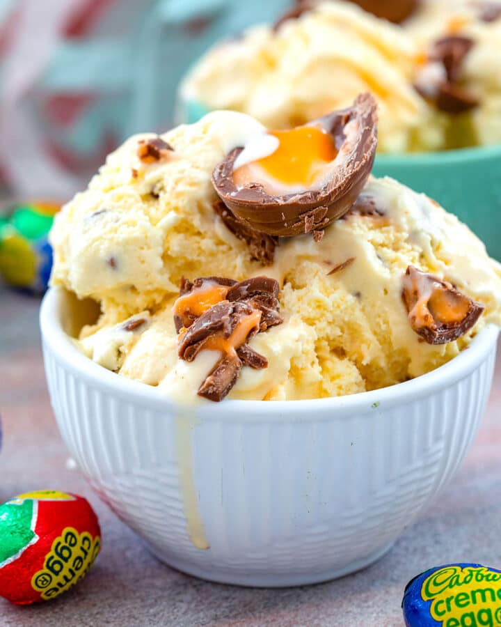 Head-on view of a bowl of Cadbury Creme Egg ice cream topped with a creme egg with wrapped candy all around and second bowl of ice cream in background