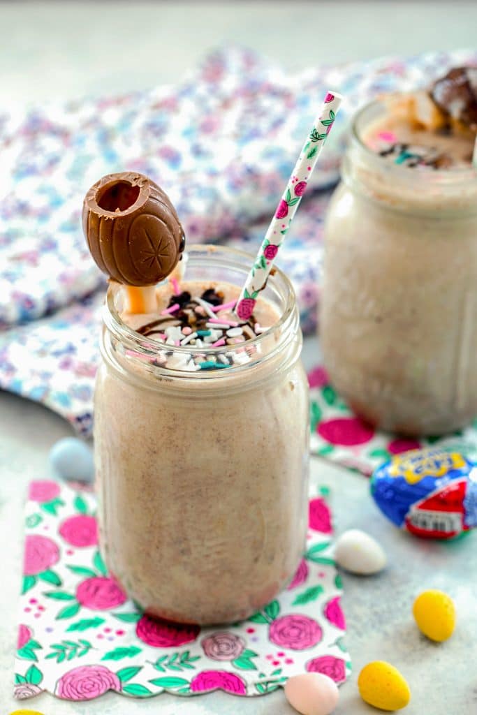 Head-on view from a distance of Cadbury Egg Milkshake with sprinkles, Cadbury Egg, and a straw surrounded by Cadbury Mini Eggs with a second milkshake in the background.