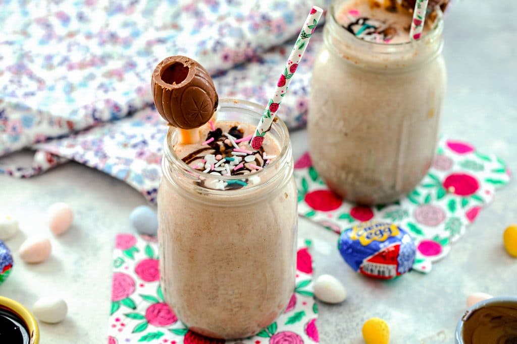 Landscape view of two Cadbury Cream Egg milkshakes with sprinkles, Cadbury Cream Egg garnish, and straw, on flowered napkins and surrounded by Cadbury Mini Eggs