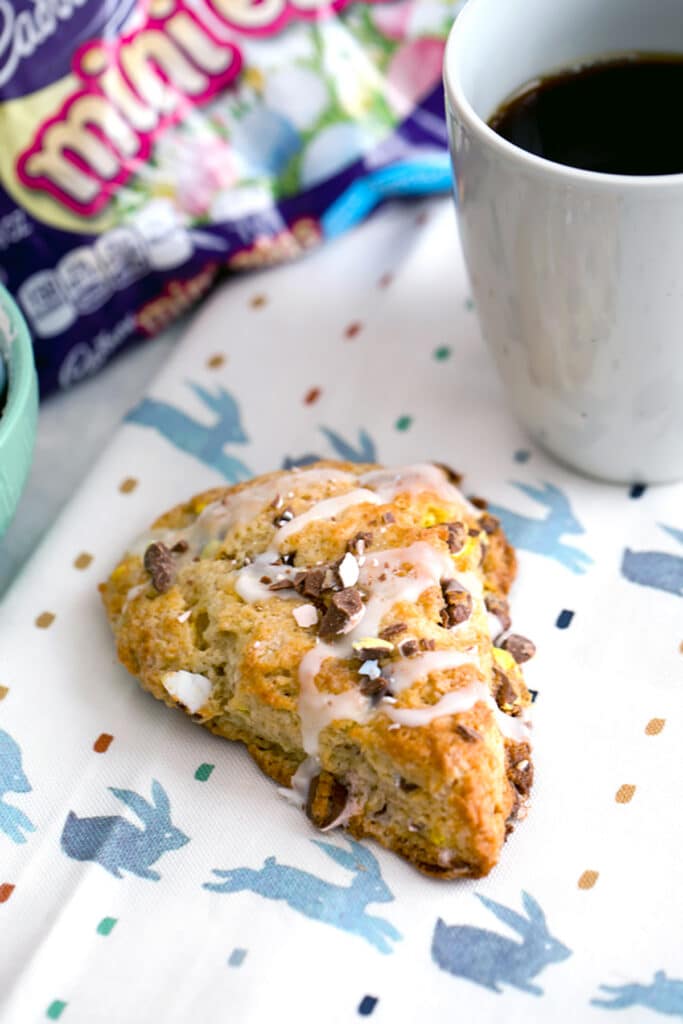 Overhead view from a distance of Cadbury Mini Egg scone on a bunny towel with bag of mini eggs and cup of coffee in the background