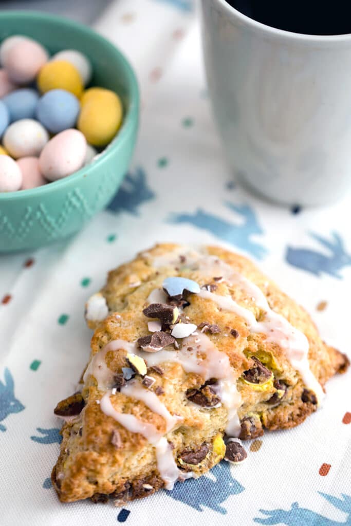 Overhead view of a Cadbury Mini Egg scone on a bunny towel with a bowl of mini eggs and a cup of coffee in the background and recipe title at top.